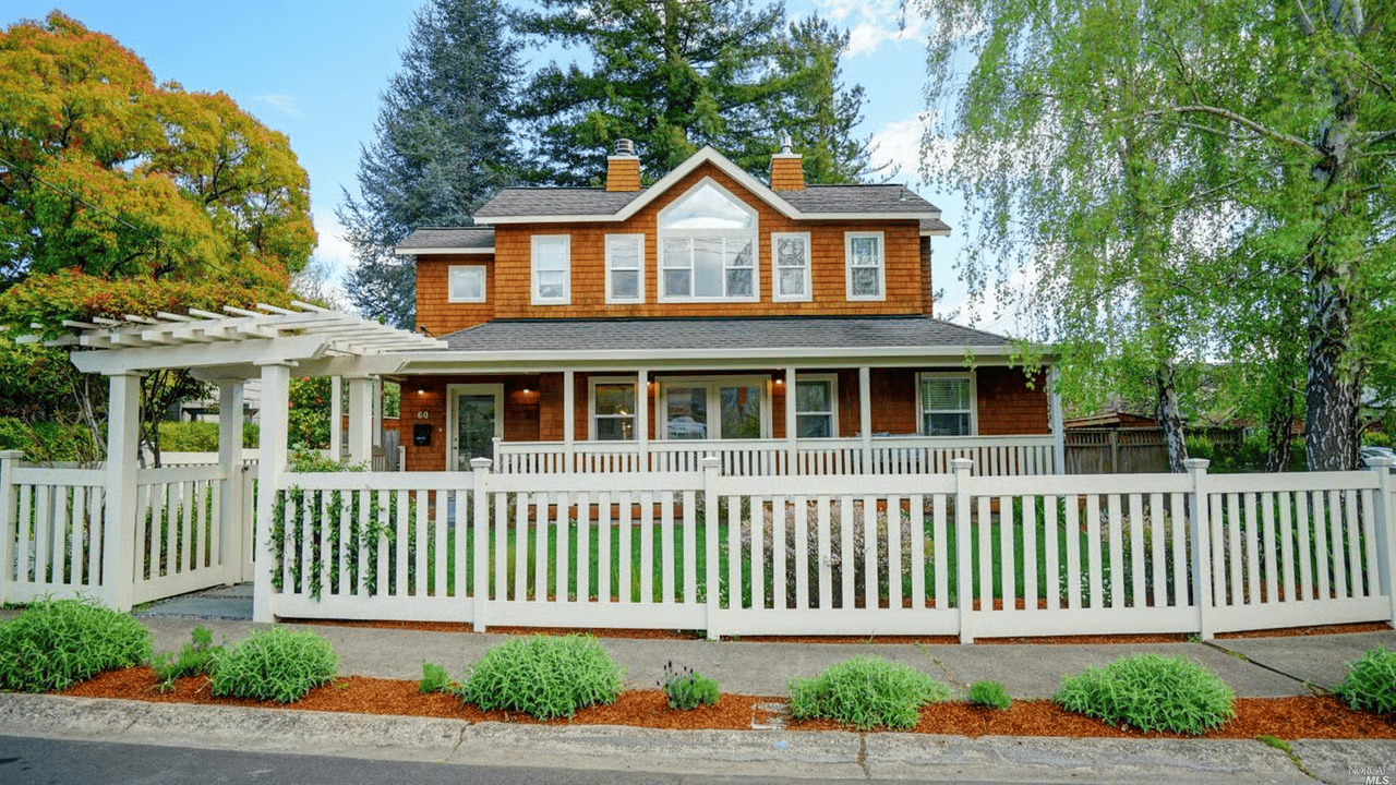 Two-story cedar-shingle house behind a white picket fence, with a covered porch and pergola.