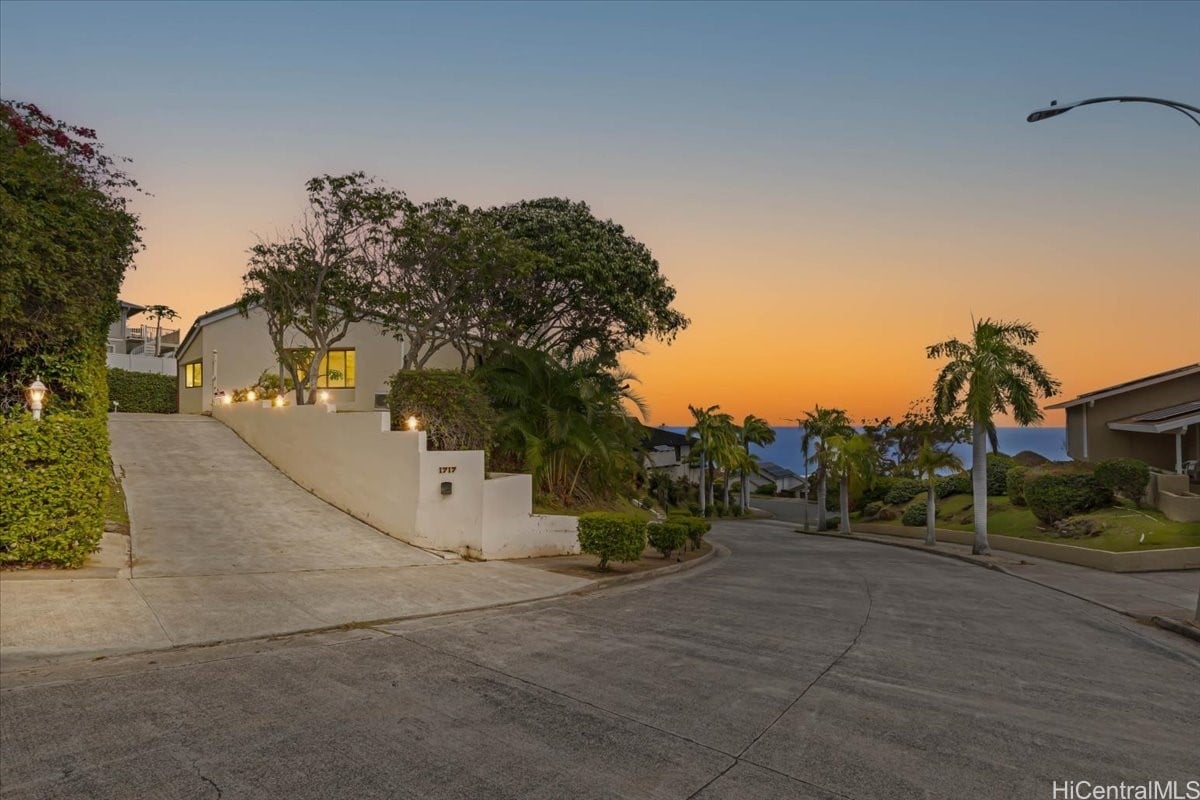 Modern home with a sloped driveway and tropical landscaping, overlooking the ocean at dusk.