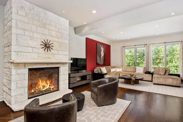 Living room with a lit stone fireplace, red accent wall, and large windows overlooking greenery.