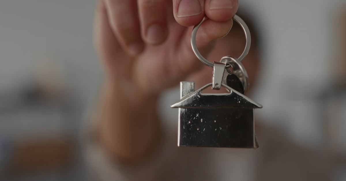 A close-up of a person's hand holding a key ring with keys and a small, metal house as a keychain toward the viewer.
