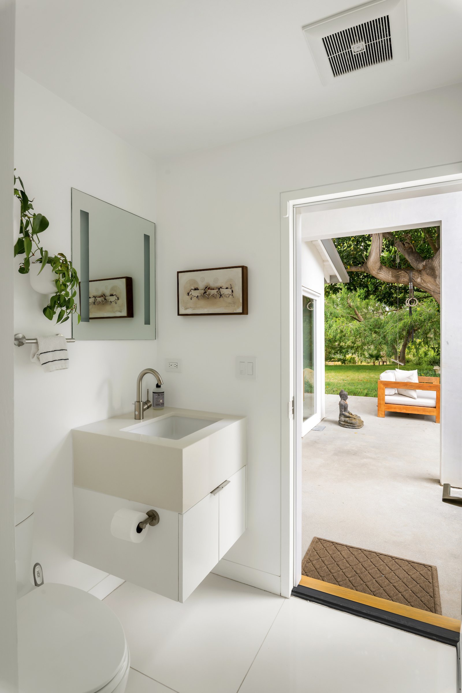 White powder room with a floating vanity opens onto a covered patio and leafy garden.