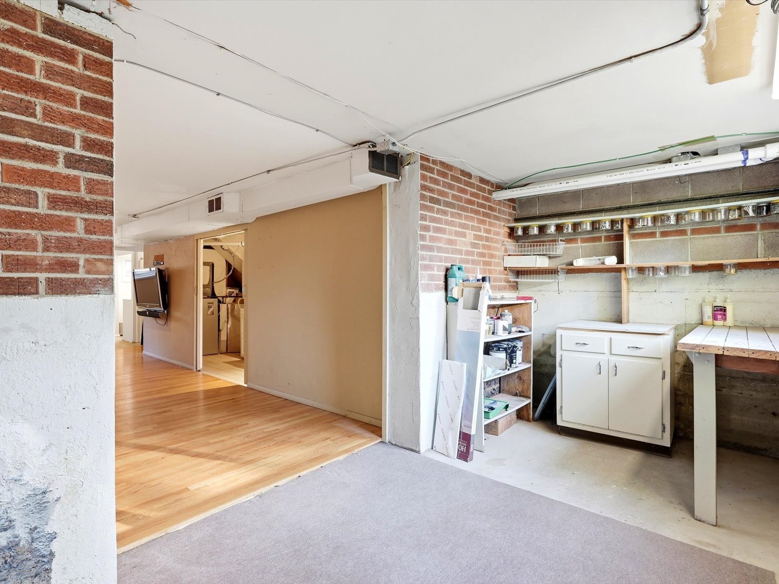 Garage workshop with brick walls, open shelves, a workbench, and white cabinets beside a wood-floored room.