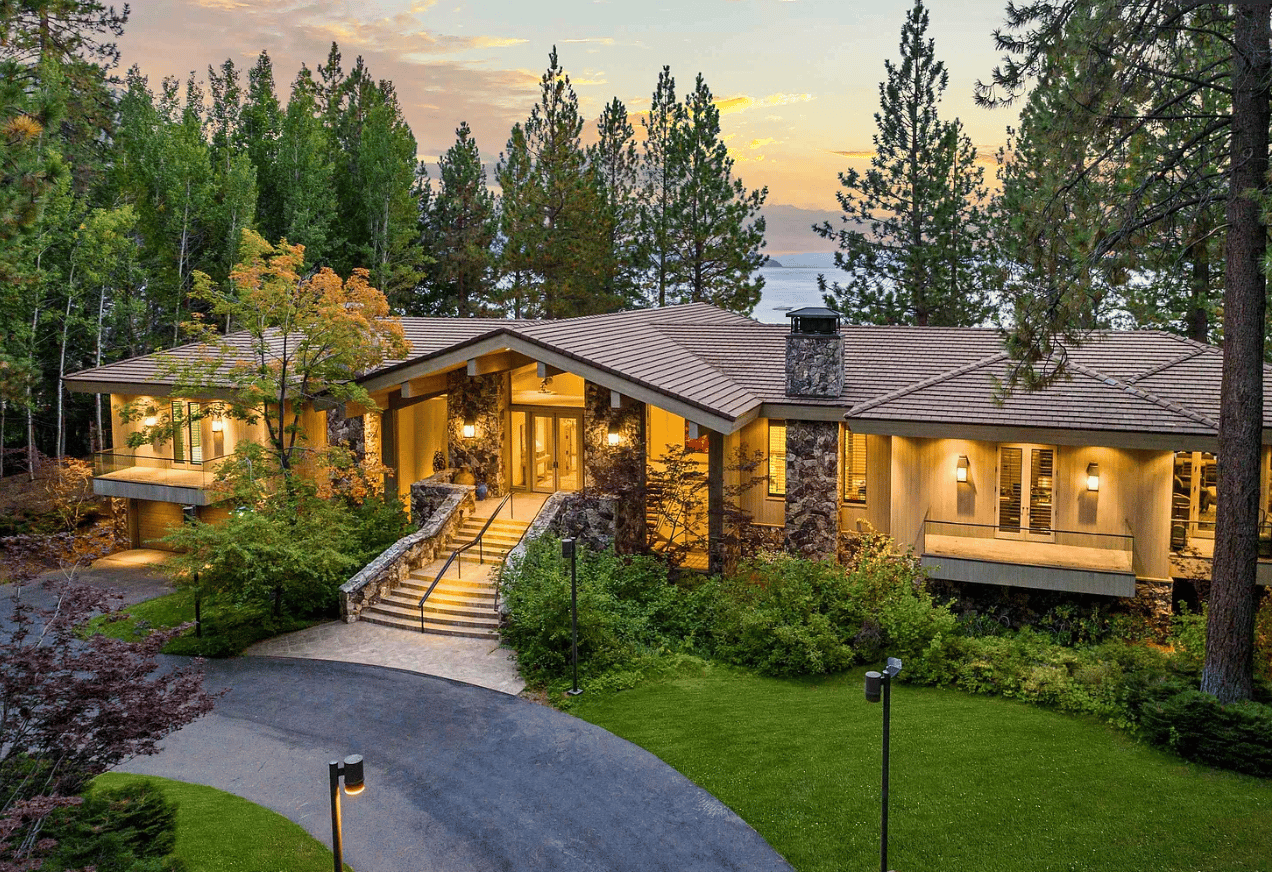 Modern stone-and-wood lakefront home with a curved driveway and Lake Tahoe visible through the trees at dusk.
