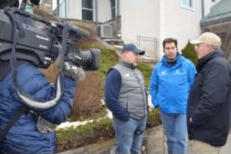Four men gather outside a house as a camera operator films them.