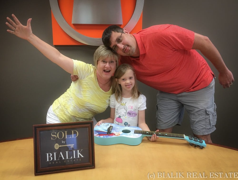 A family of three poses behind a guitar beside a Bialik Real Estate sold sign.