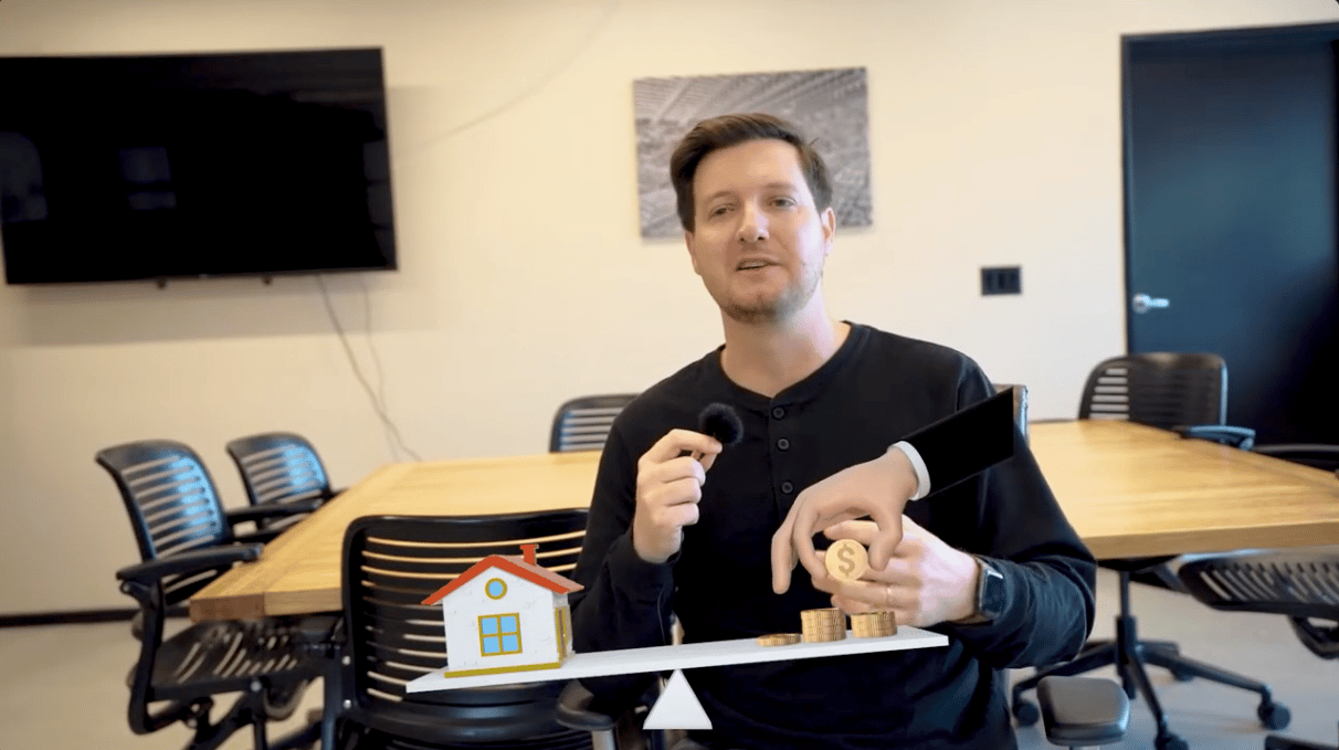 Man speaking in a conference room beside a model house and coin stacks.