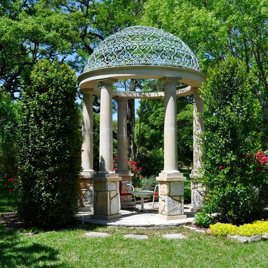 Garden gazebo with a white filigree dome and stone-based columns surrounded by lush greenery and flowers.