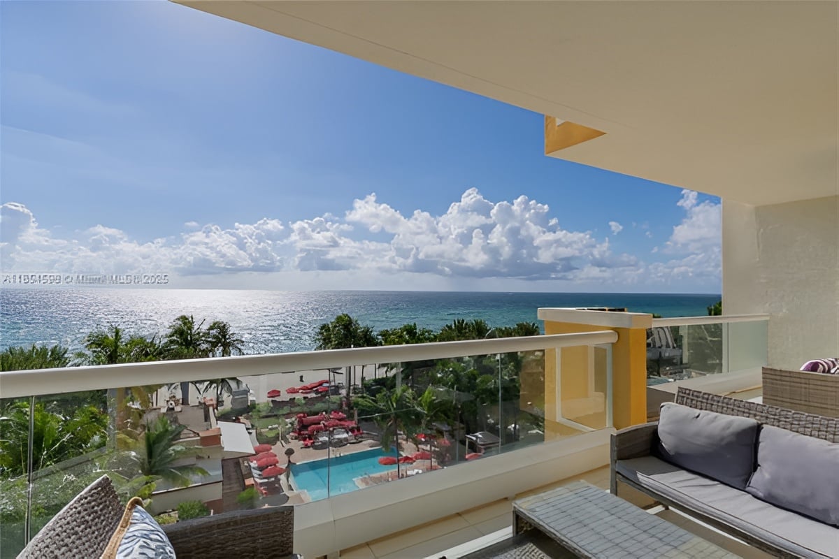 Furnished balcony overlooks the ocean and a resort pool with red loungers.