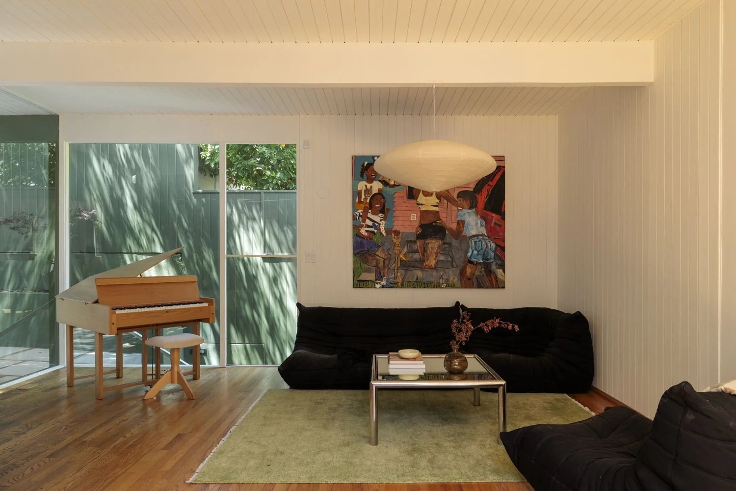 The open living room of the Smith Residence with a white board ceiling, wood floors, a low black sofa, and a wall of glass looking out to green siding and trees.