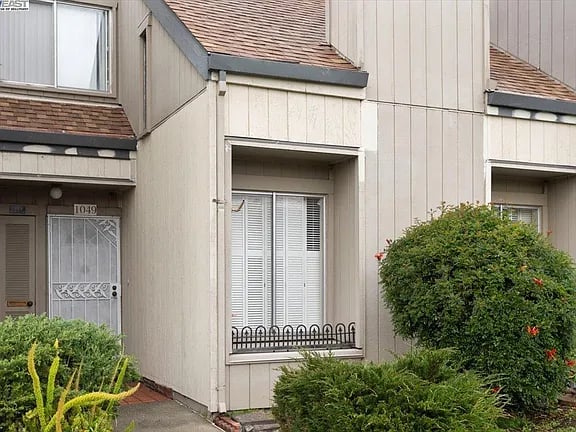 Beige residence entrance marked 1049, with white blinds and flowering shrubs.