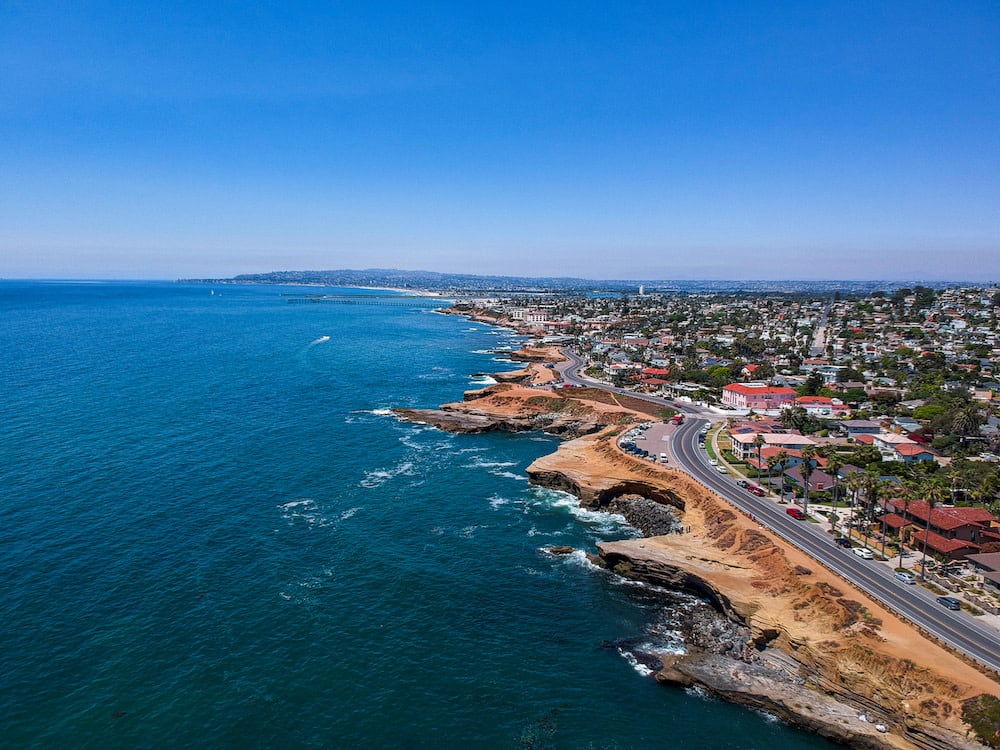 Aerial coastline with rocky cliffs, a winding coastal road, and homes beside the ocean.
