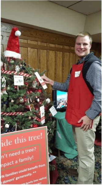 Man in a red apron beside a decorated Christmas tree at an indoor festival.