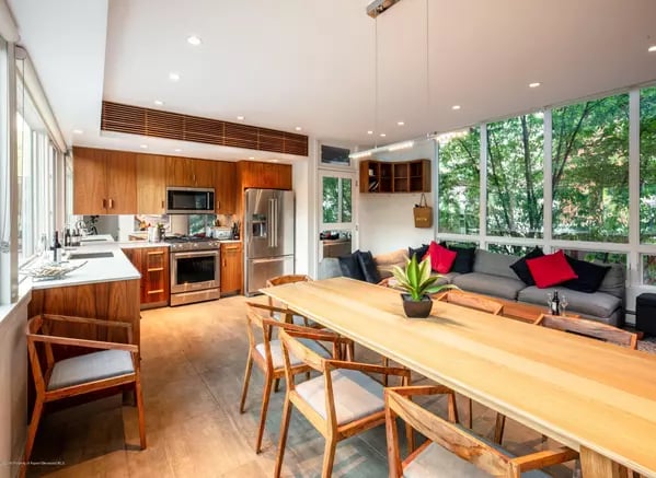 Open-plan kitchen and dining area beside a dark sectional and floor-to-ceiling windows overlooking trees.