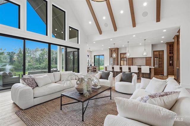 Open-plan living room with white sofas, vaulted wood-beam ceiling, and large windows overlooking greenery.