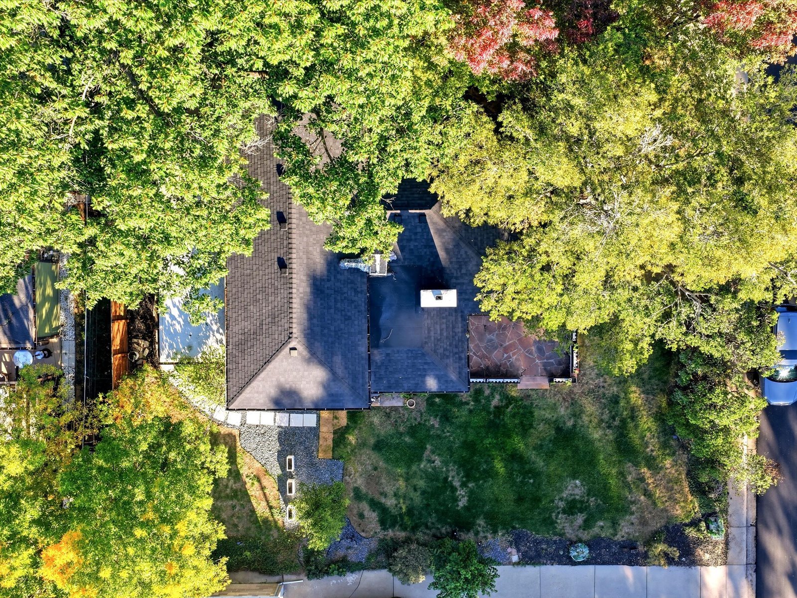 Aerial view of a tree-shaded home with a dark roof, stone patio, and front lawn.