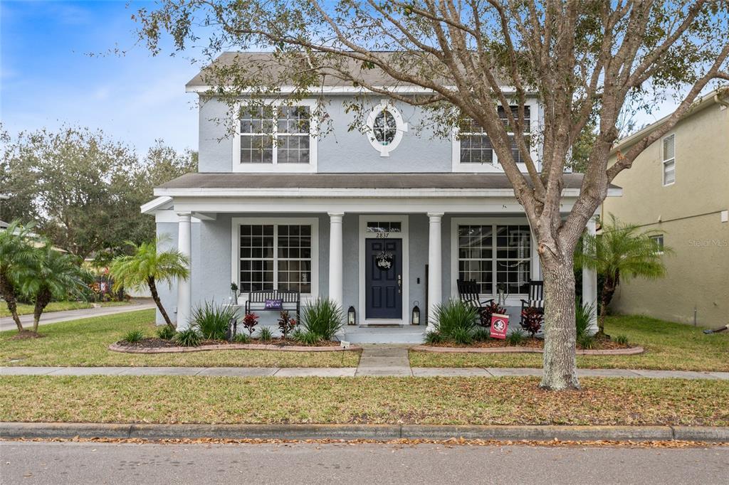 Two-story light-gray house with a covered porch, dark blue door, and large front-yard tree.