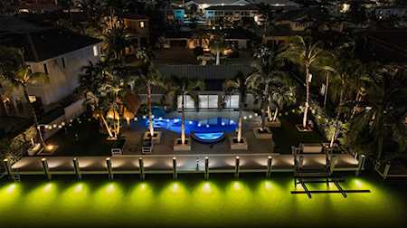 Waterfront home with a lit pool and dock over glowing water at night.