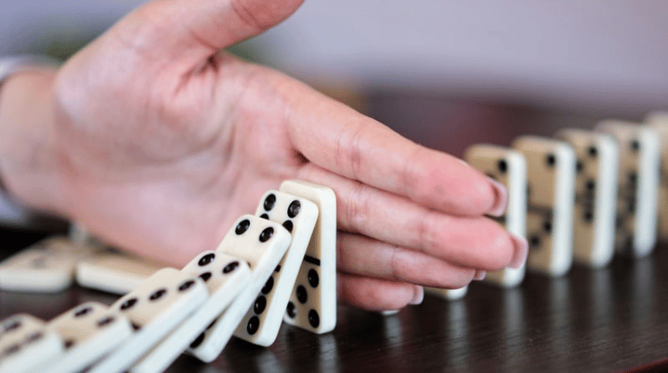 A hand stops a line of falling dominoes on a dark tabletop.