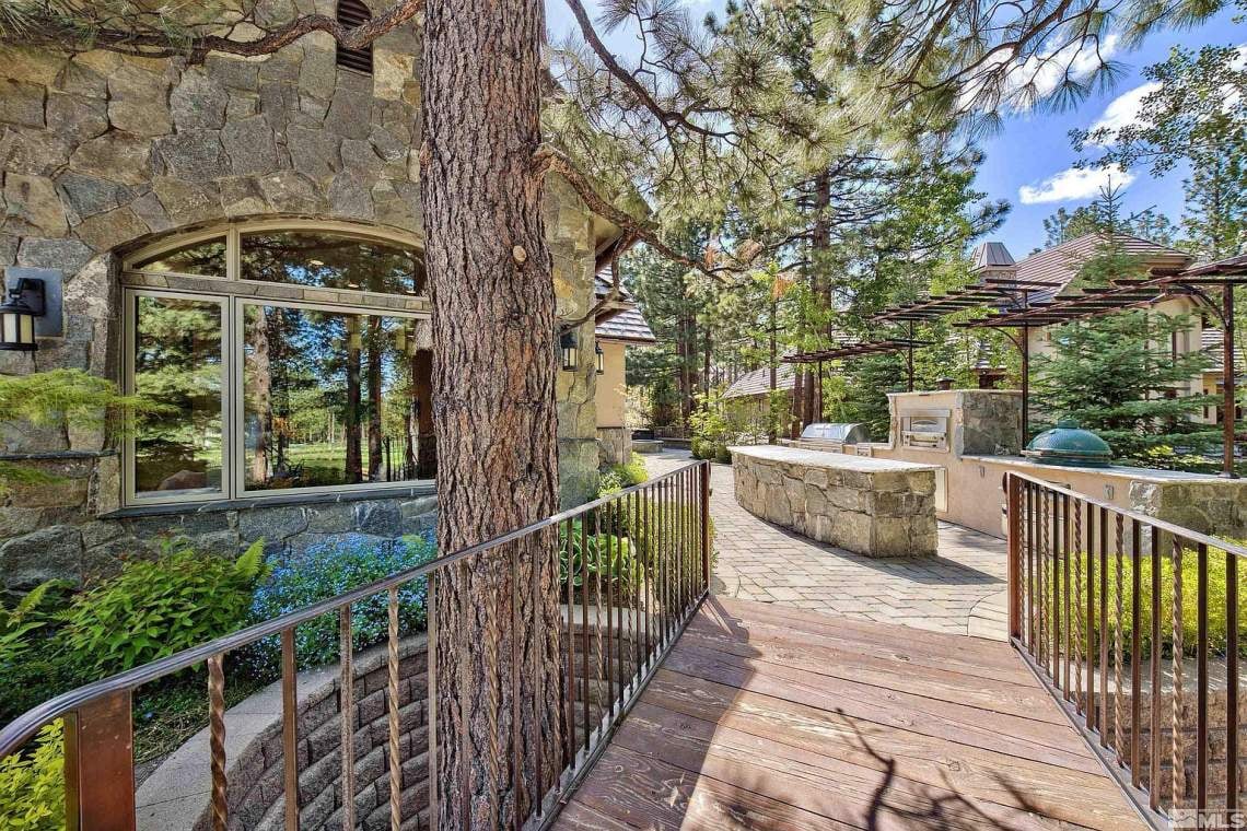 Stone home patio with a deck walkway, built-in grill, pergola, and mature pine trees.