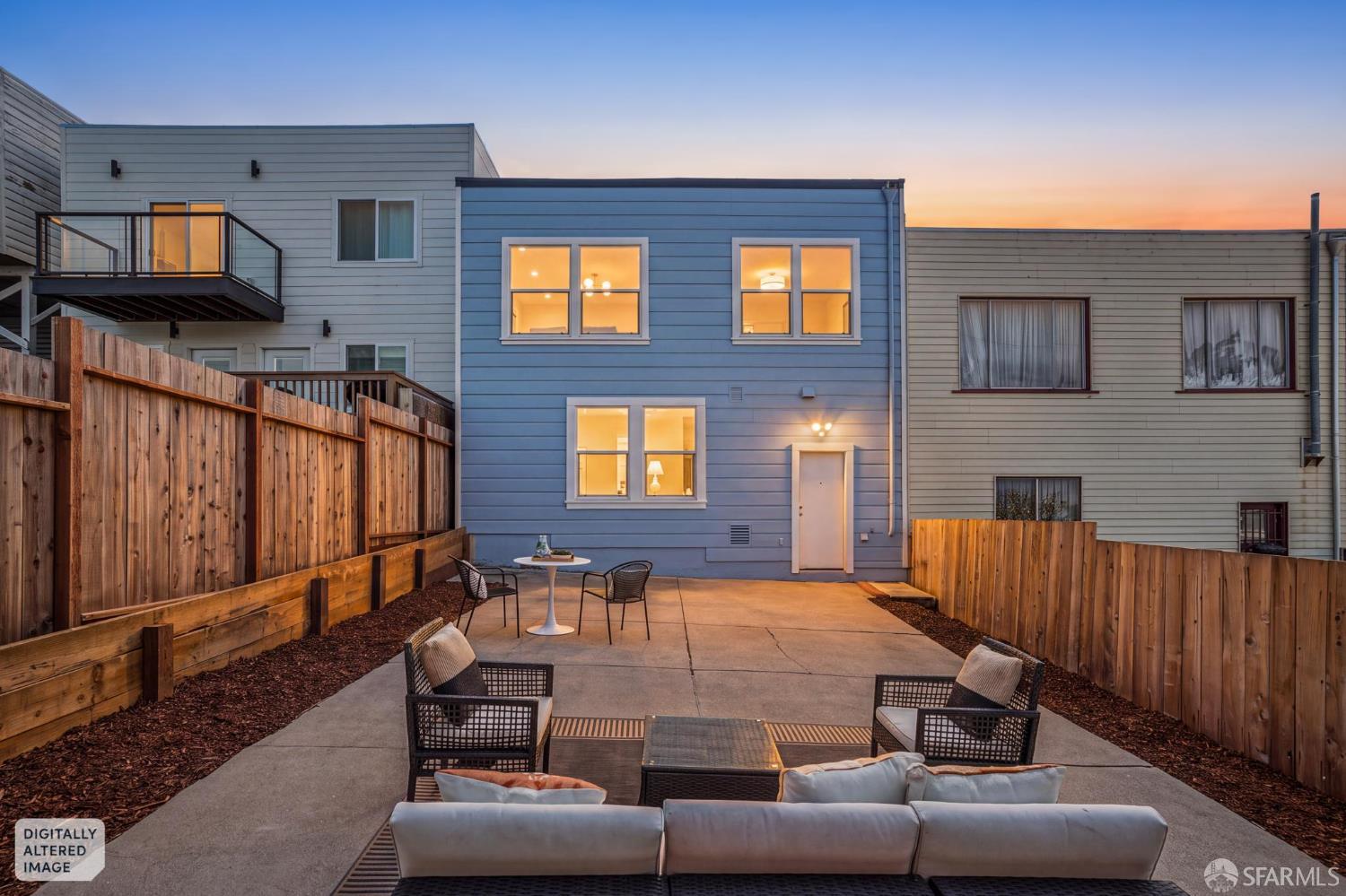 Modern home’s rear patio at dusk, with fenced concrete seating area and warmly lit windows.