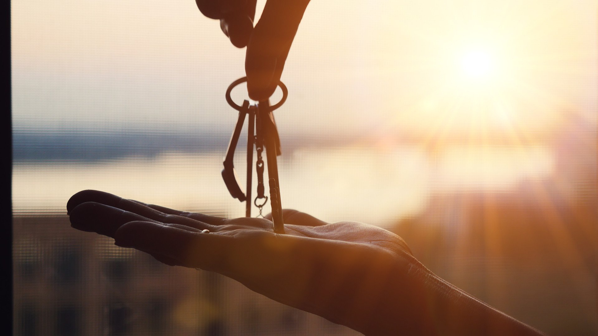A hand offers house keys to another open palm in warm backlight.