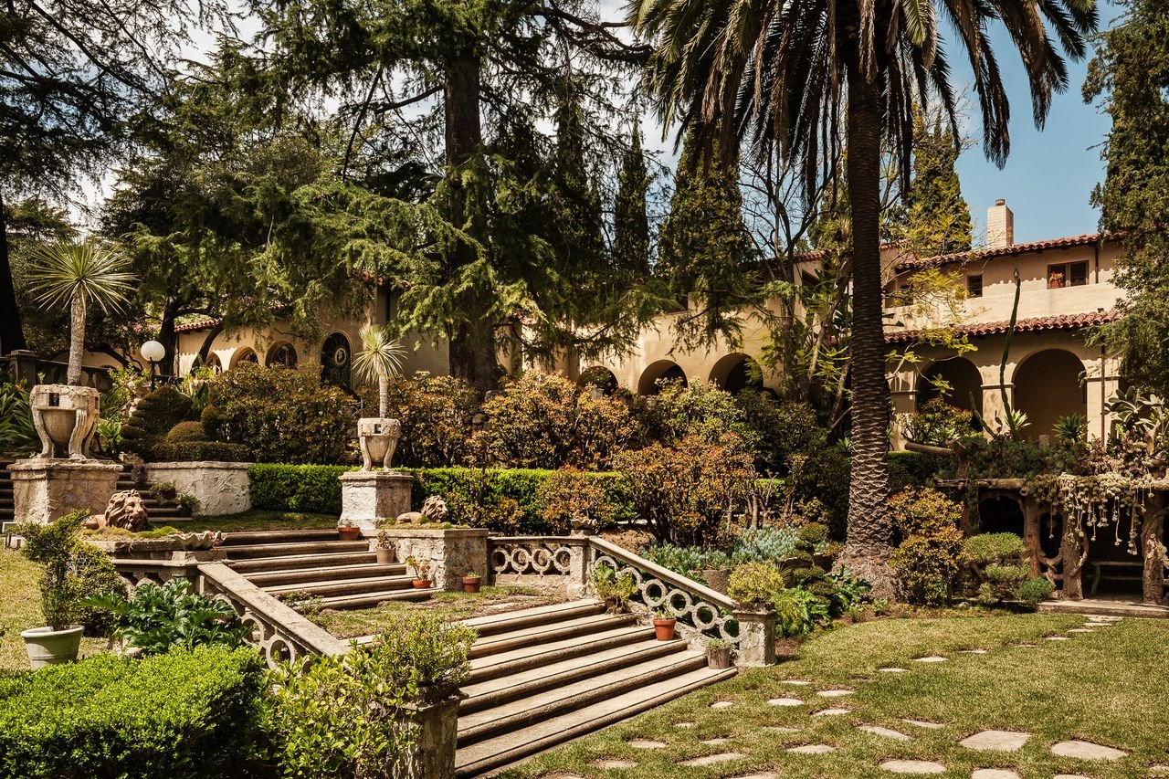 Mediterranean-style home behind a landscaped courtyard with stone steps, arched arcades, and mature trees.