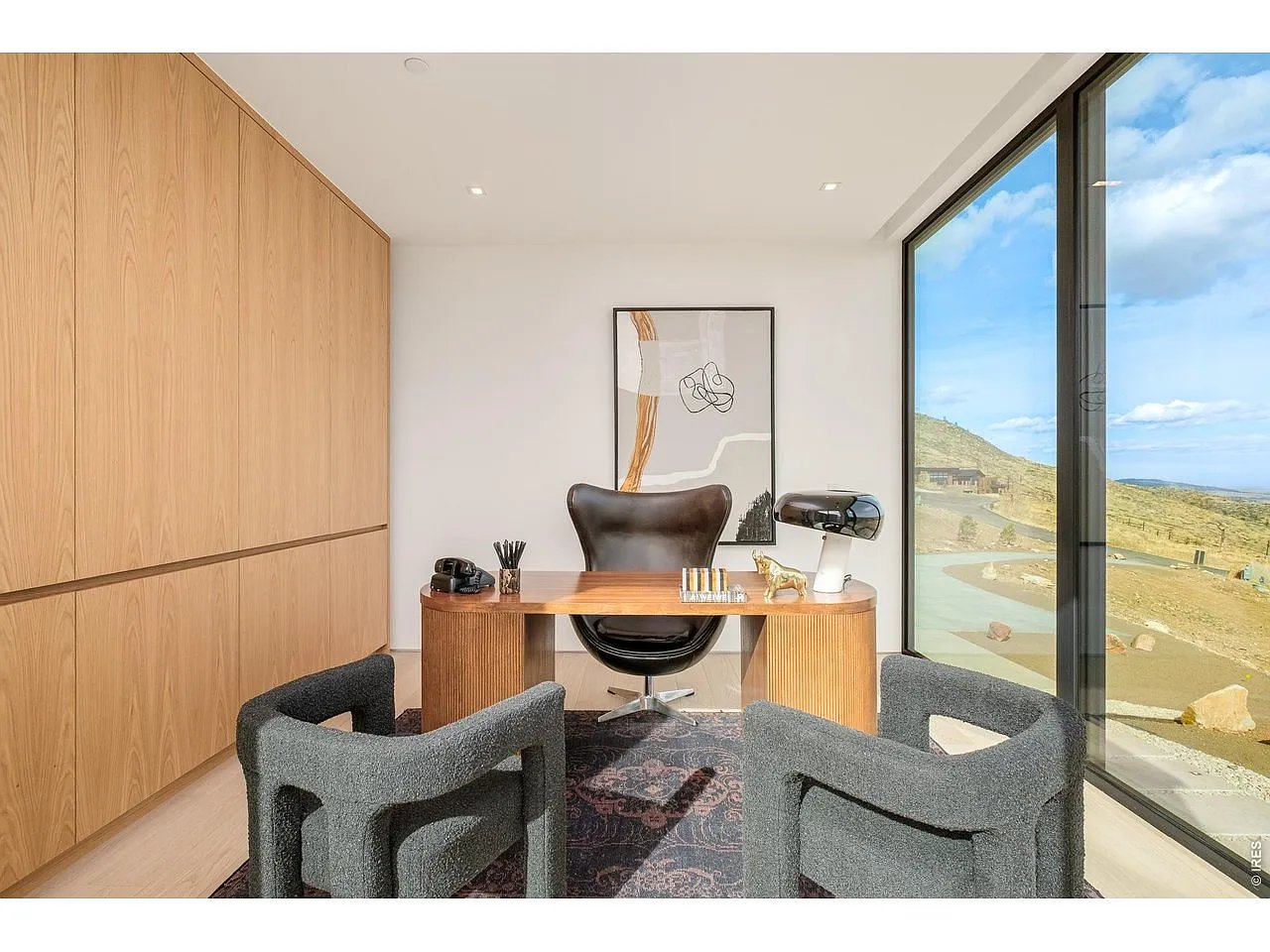 Home office with a curved wood desk, gray guest chairs, and floor-to-ceiling windows overlooking a hillside.