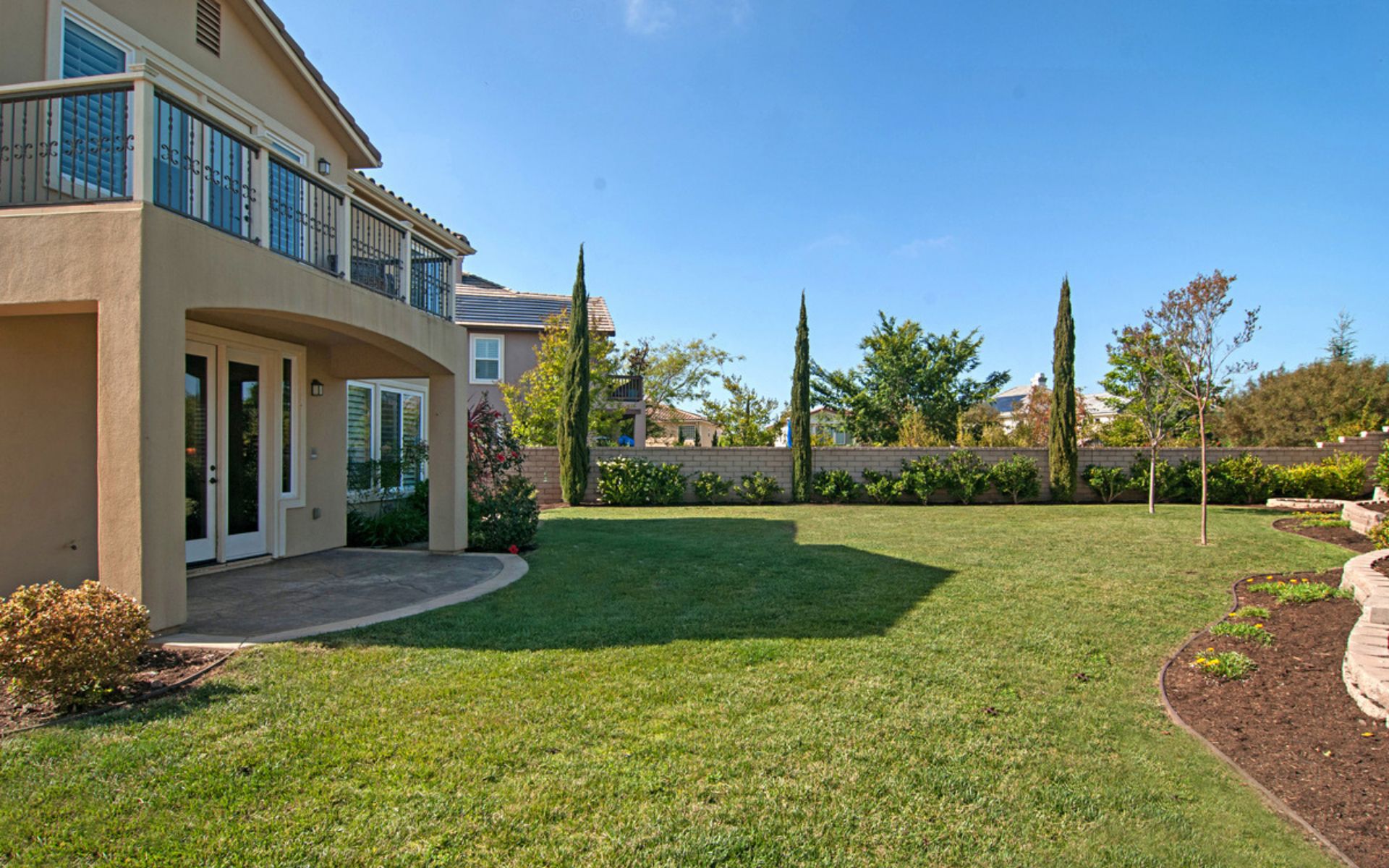 Home backyard with a broad lawn, curved garden beds, covered patio, and upper-level balcony.