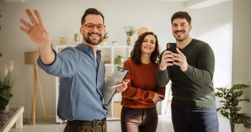 A real estate agent waves beside a couple in a bright living room.
