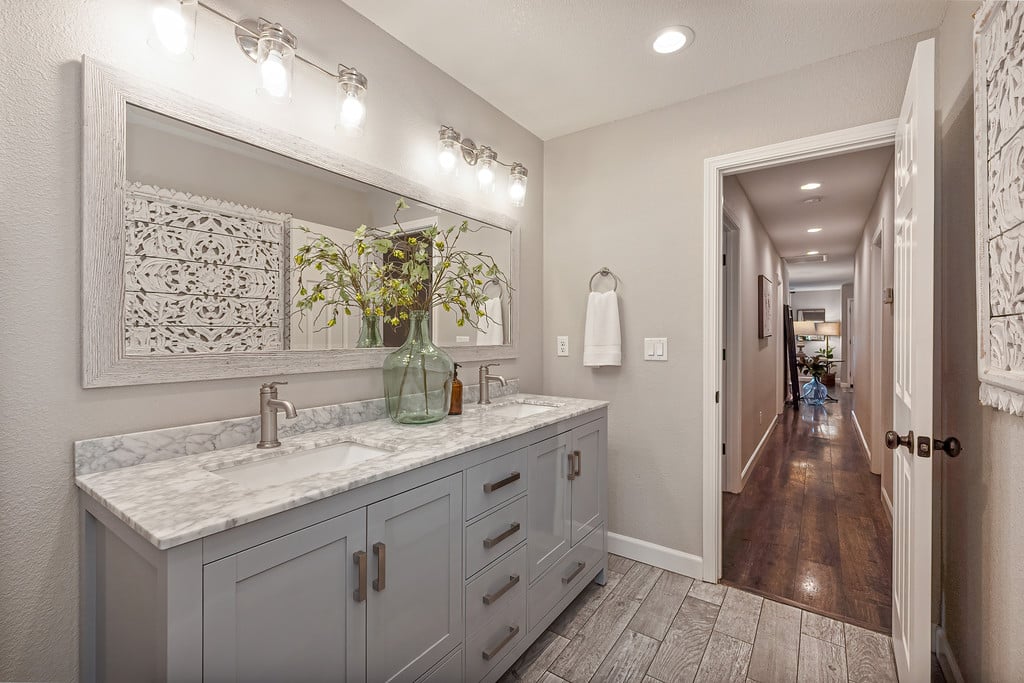 Bathroom vanity with gray cabinetry, marble-look countertop, and large mirror beside an open hallway.