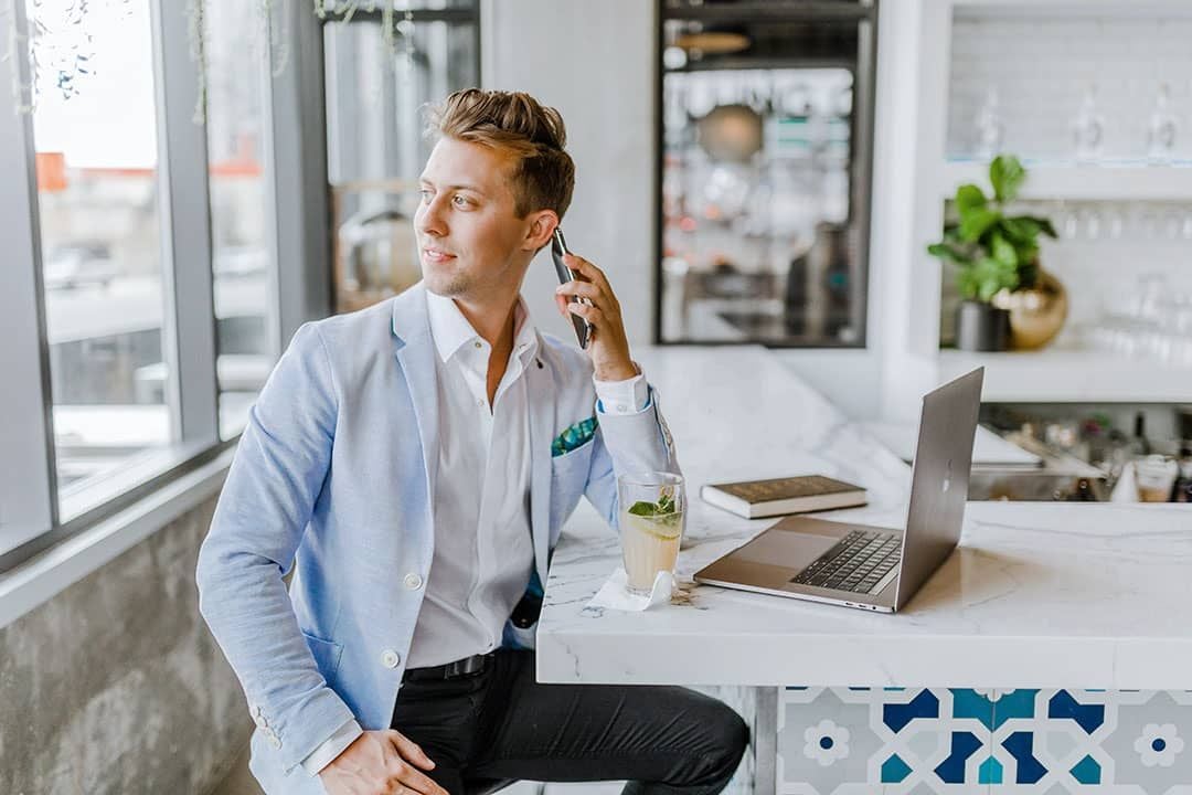 Man in a light blue blazer talks on his phone beside an open laptop at a marble counter.