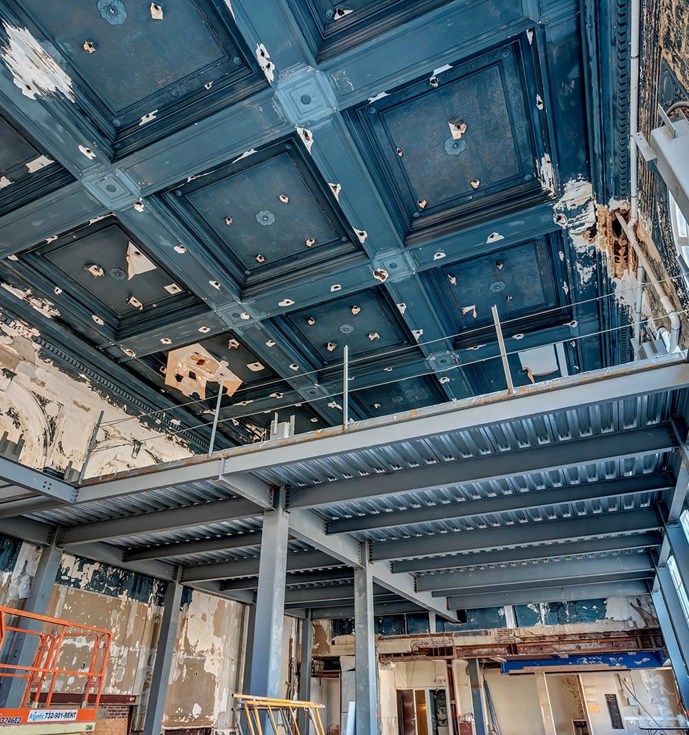 Interior under construction with exposed steel framing beneath a weathered blue coffered ceiling.