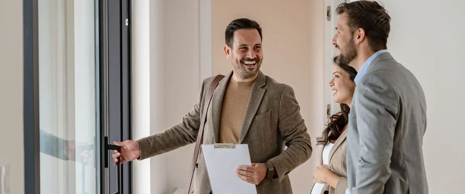 A smiling real estate agent opens a glass door for a couple touring a home.