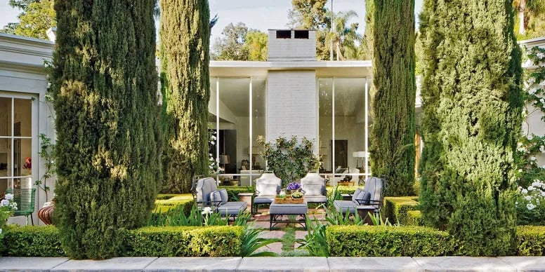 Modern white house behind a formal garden courtyard with tall cypress trees and outdoor seating.