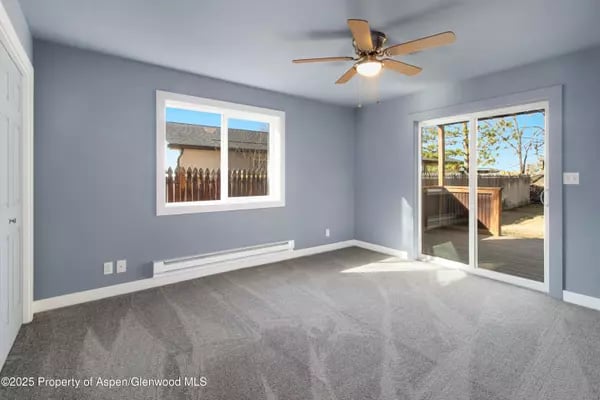 Empty gray room with carpet, a ceiling fan, and sliding glass doors to a fenced outdoor area.