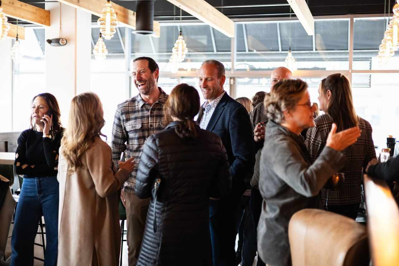 Adults mingle and converse beneath timber beams and pendant lights at a gathering.