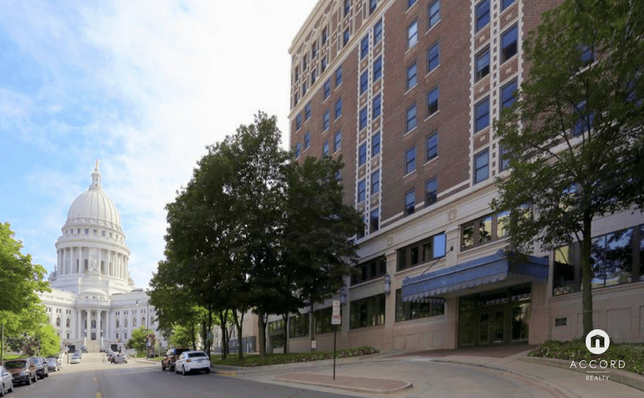 Brick urban condominium beside a tree-lined street, with the domed Wisconsin State Capitol in the background.