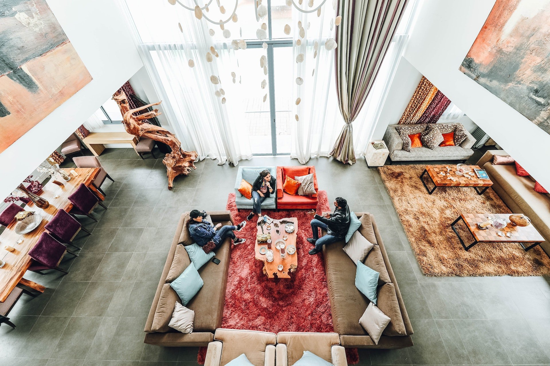 Open-plan living room with floor-to-ceiling windows, red rugs, and people seated around the coffee table.