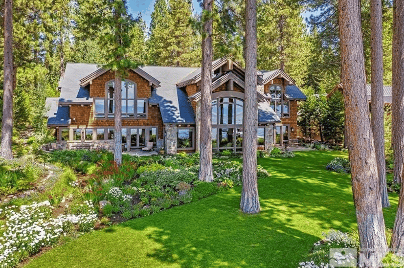Rustic stone-and-timber home nestled among pines, with a broad green lawn and landscaped flower beds.