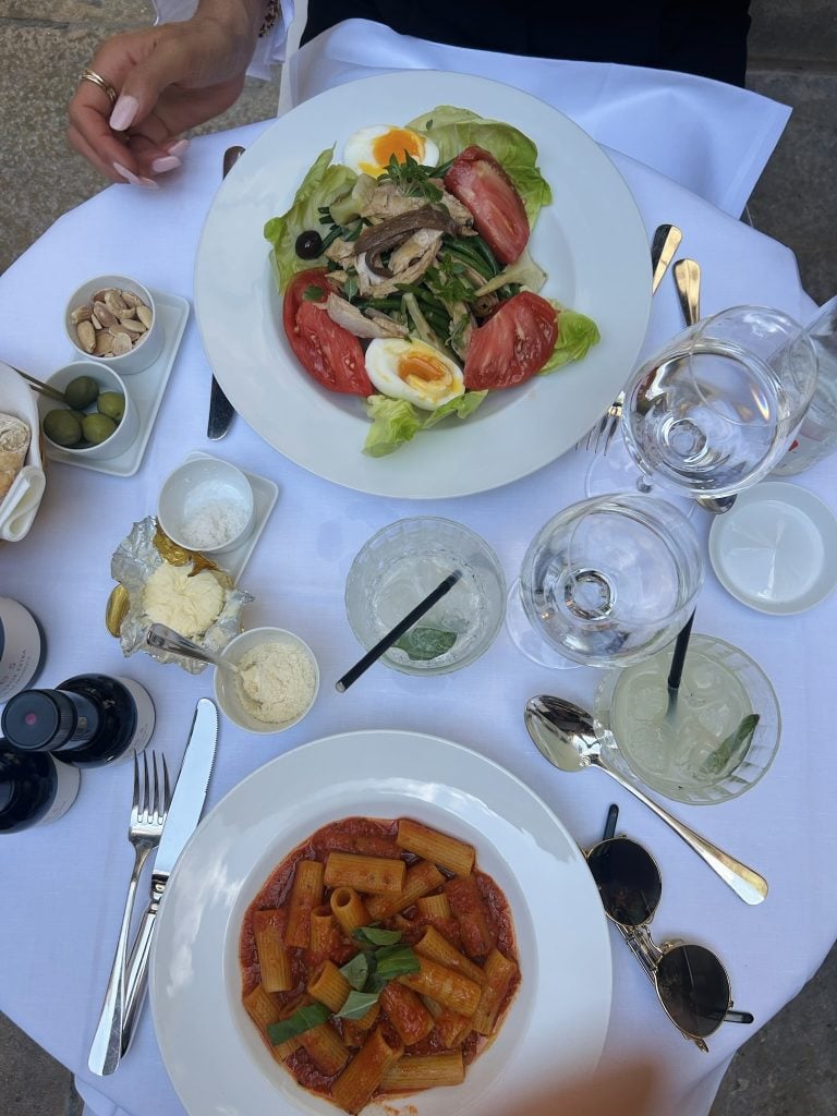 Outdoor table set with tomato and egg salad, rigatoni in tomato sauce, olives, and drinks.