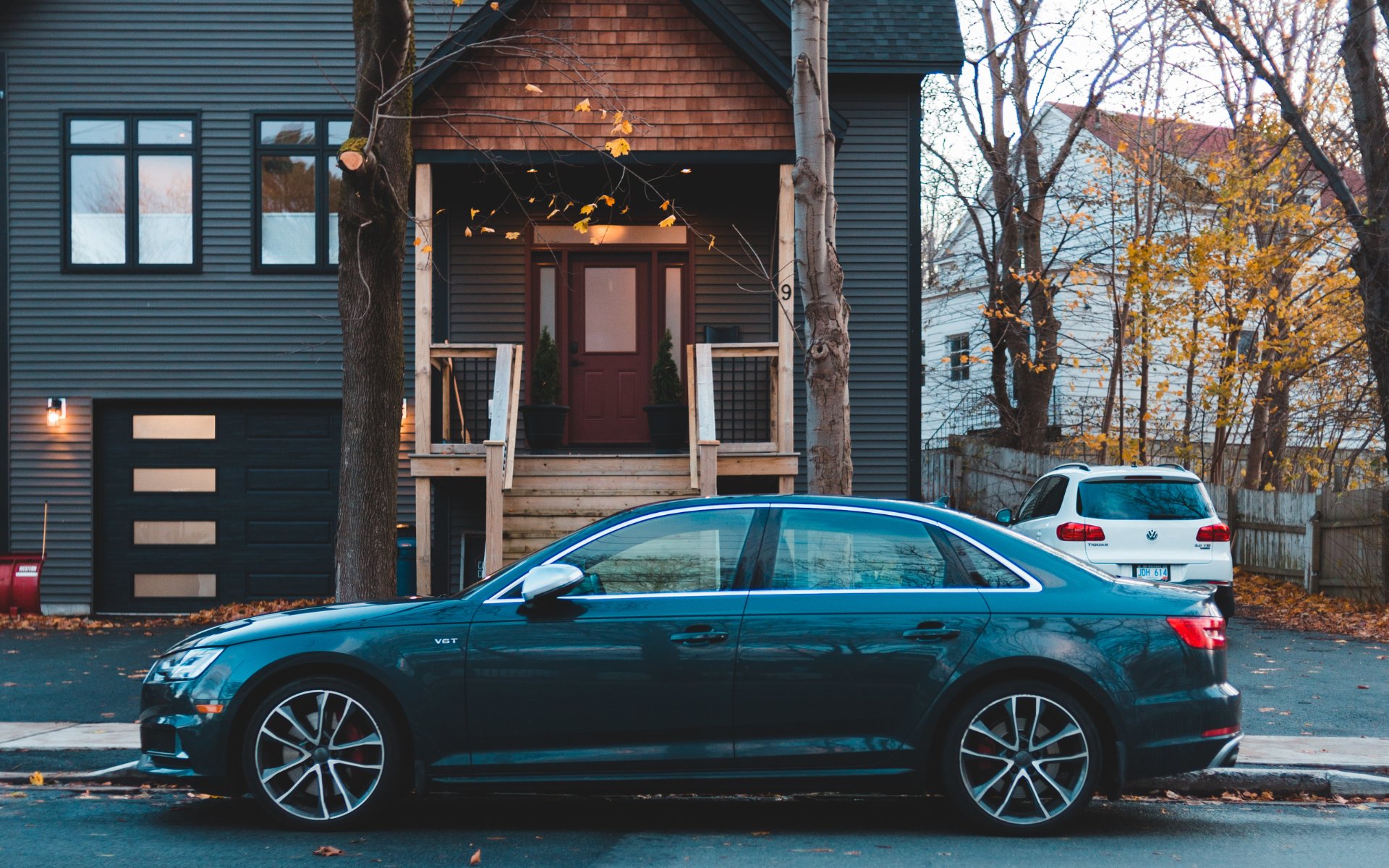 Two-story blue-sided house with a raised porch and garage on a compact city lot, partly obscured by parked cars.