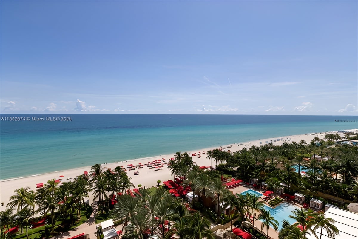 Beachfront resort with turquoise water, red beach umbrellas, palm trees, and a swimming pool.