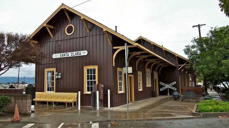 Historic Santa Clara Caltrain station building with a &ldquo;Santa Clara&rdquo; sign, covered platform, and wooden benches on an overcast day.