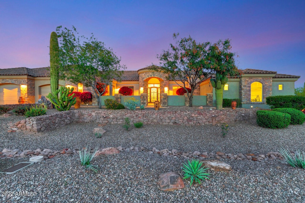 Single-story stone-and-stucco home with a lit entry, stone wall, and desert landscaping at dusk.