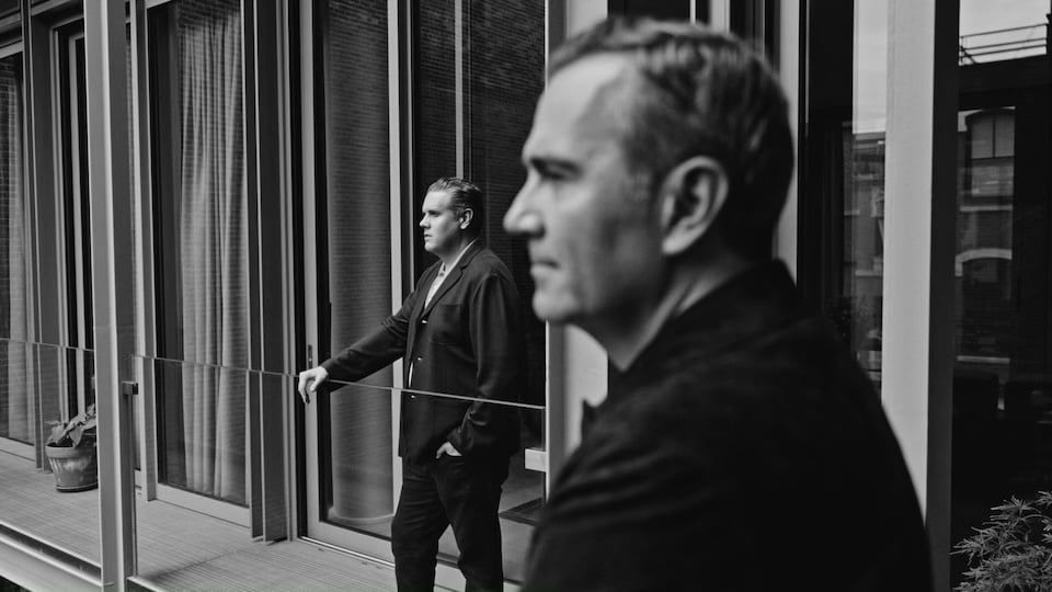 Two men in dark suits on a modern glass-fronted balcony.