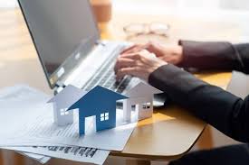 Hands typing on a laptop beside model houses and housing paperwork.