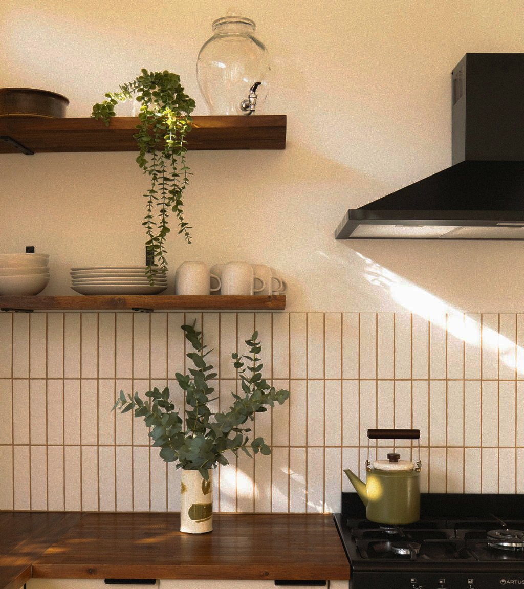 Kitchen with cream tile backsplash, wooden shelves, trailing plant, and green kettle in sunlight.