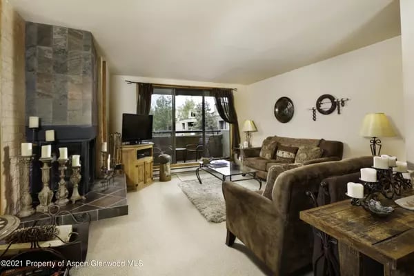 Living room with a stone fireplace, brown sofas, and large windows overlooking trees.