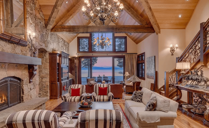 Living room with a stone fireplace, vaulted wood ceiling, and lake views through large windows.