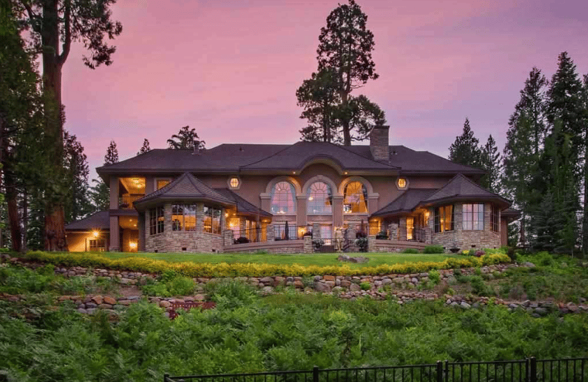 Large stone-and-wood house with arched windows, landscaped grounds, and evergreens at dusk.